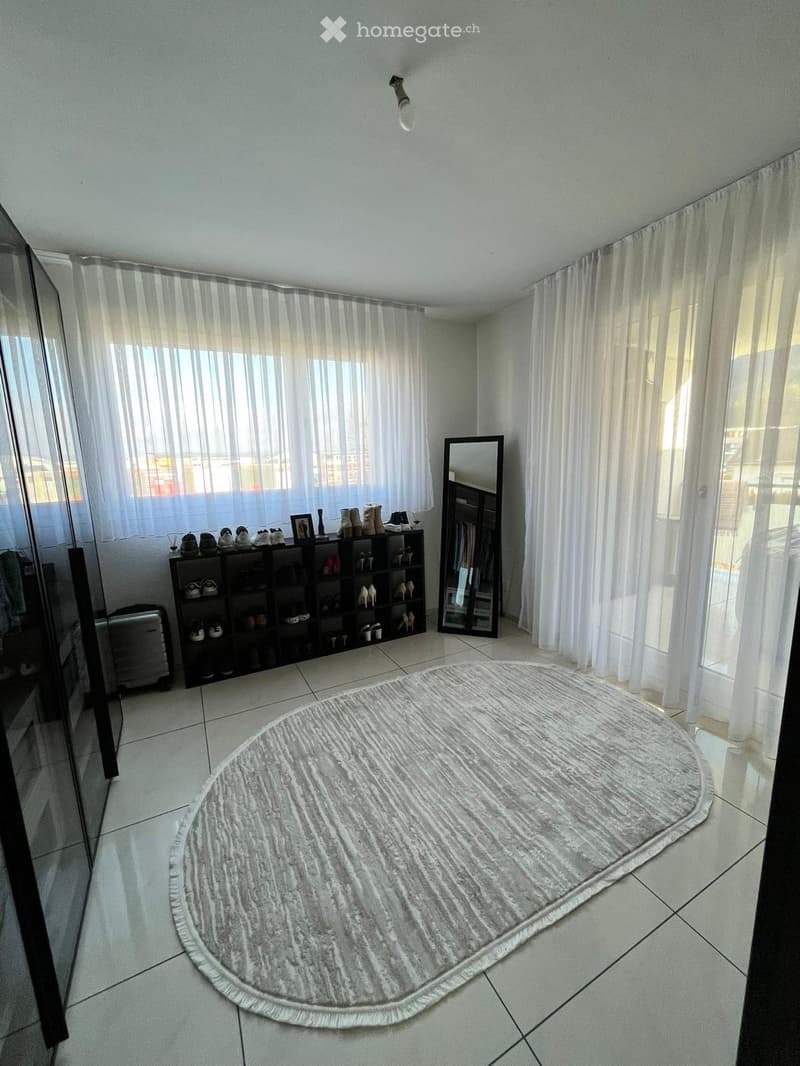 A well-lit entryway with a round grey rug, a shoe rack, a mirror, and a curtained window.