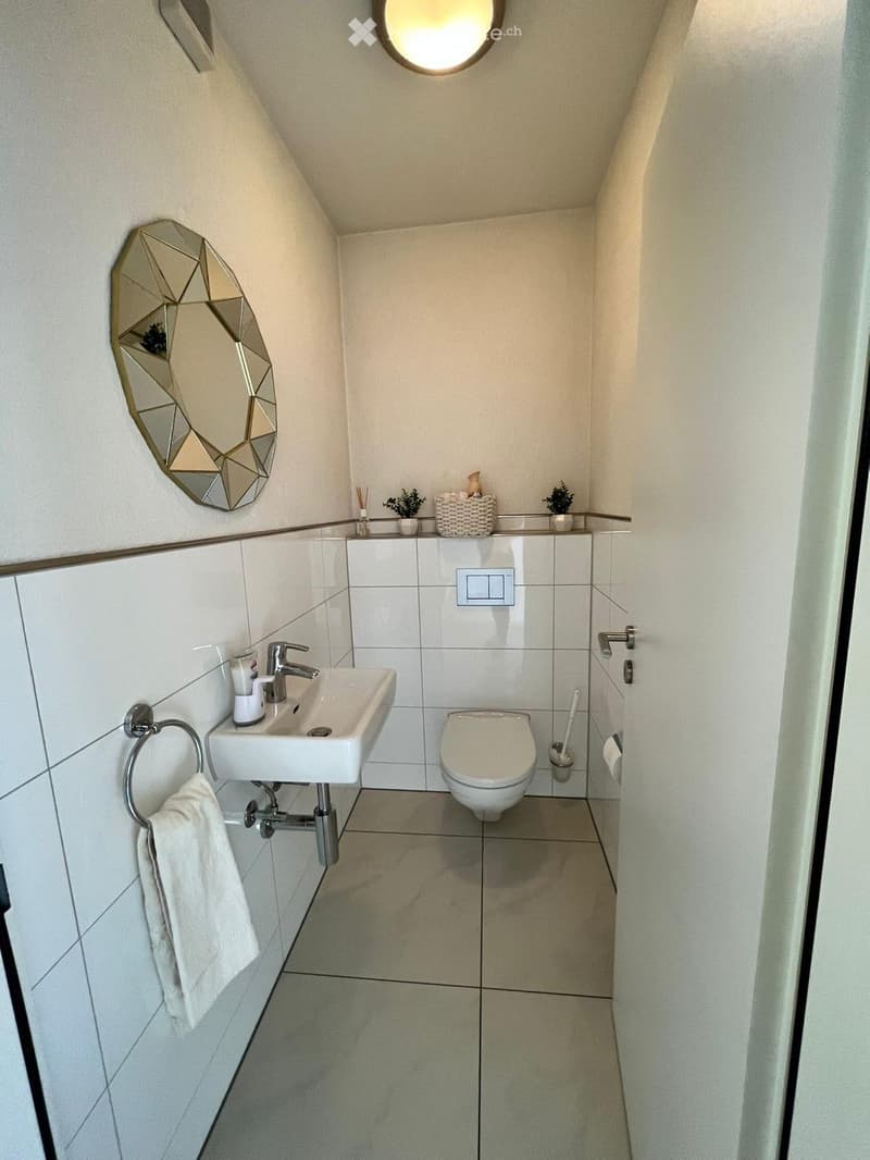 Modern bathroom with white tiles, a sink, toilet, and a decorative mirror. Includes a towel on a ring and a small shelf with decorative items.