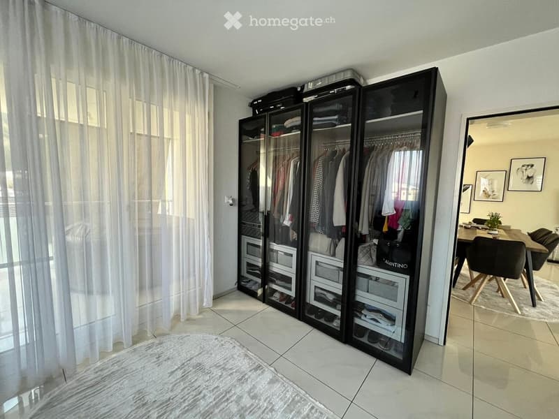 A well-lit room with a large, modern wardrobe, a dining area with a table and chairs, and a white curtain covering a window.