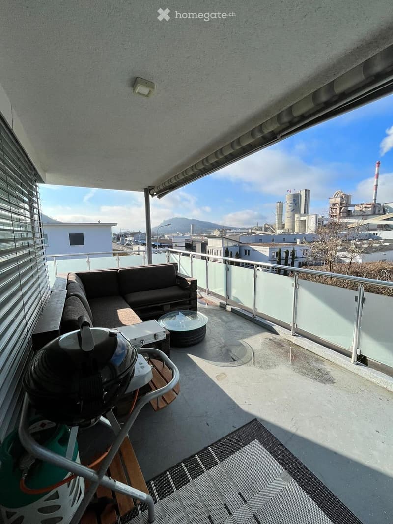 Balcony with a modern sofa, coffee table, and grill, overlooking an industrial landscape with factories and a clear blue sky.