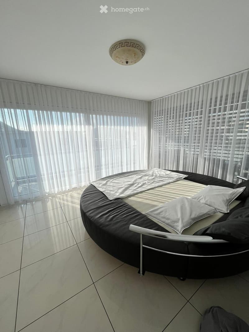 Modern bedroom with a round black bed, white pillows, and a grey blanket, large windows with sheer curtains, and a view of the city.