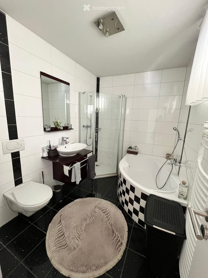 Modern bathroom with black and white tiles, a bathtub with a checkered border, a toilet, a sink with a dark wooden vanity, a shower, a round bathroom rug, and a mirror above the sink.