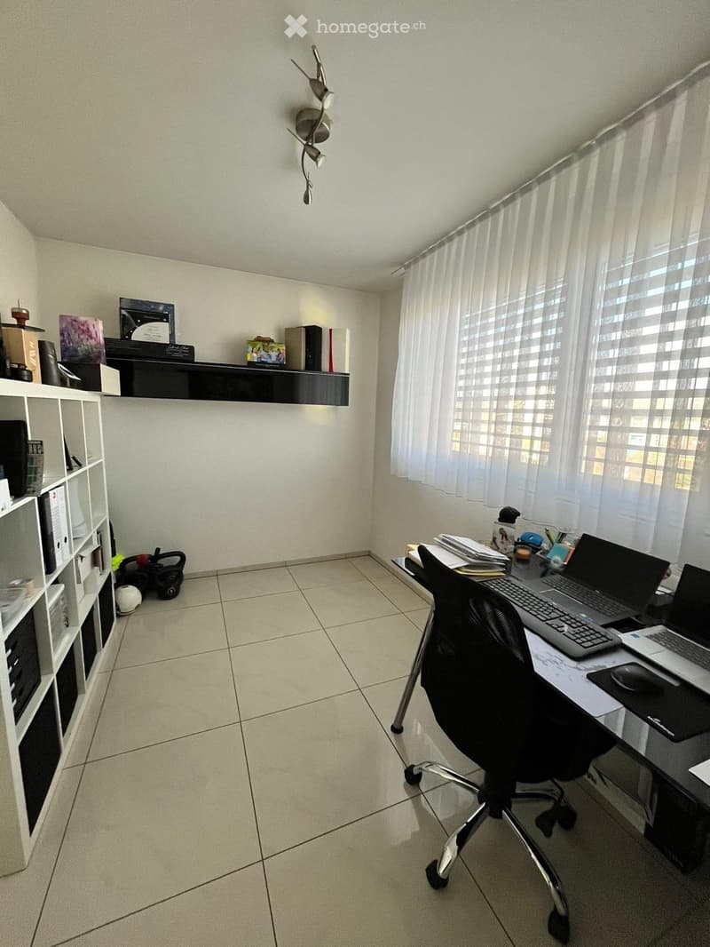 A well-lit office space with a desk, two laptops, a chair, a shelf with books and decorative items, and a window with white curtains.