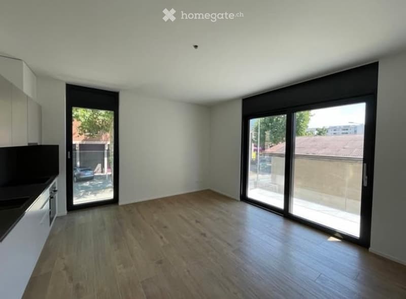 Empty room with wooden flooring, white walls, and large sliding glass doors leading to an outdoor area. Minimalist design with a small kitchen area visible on the left side.