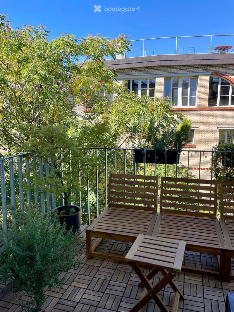 Wooden deck with two benches, a small table, and a potted plant, surrounded by greenery and a metal railing.