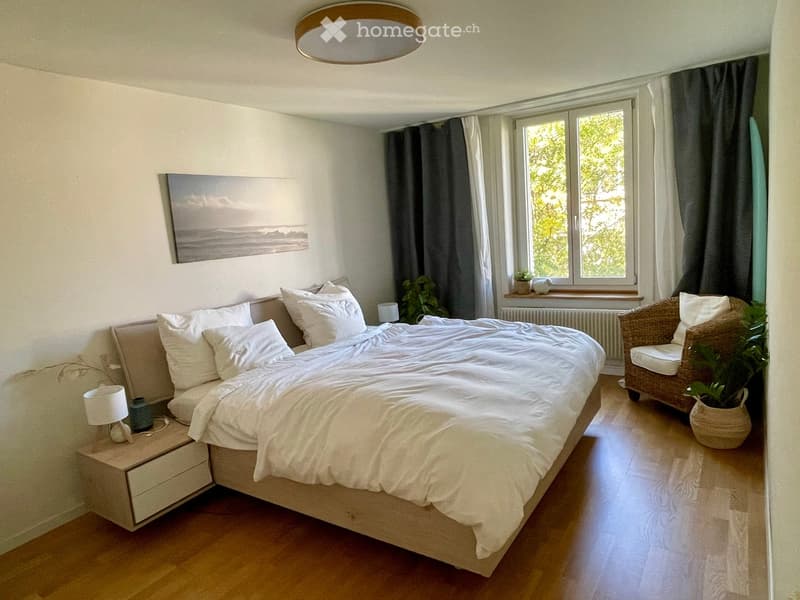 Cozy bedroom with a large bed, nightstand with lamp, wicker chair, potted plant, and a scenic picture on the wall.