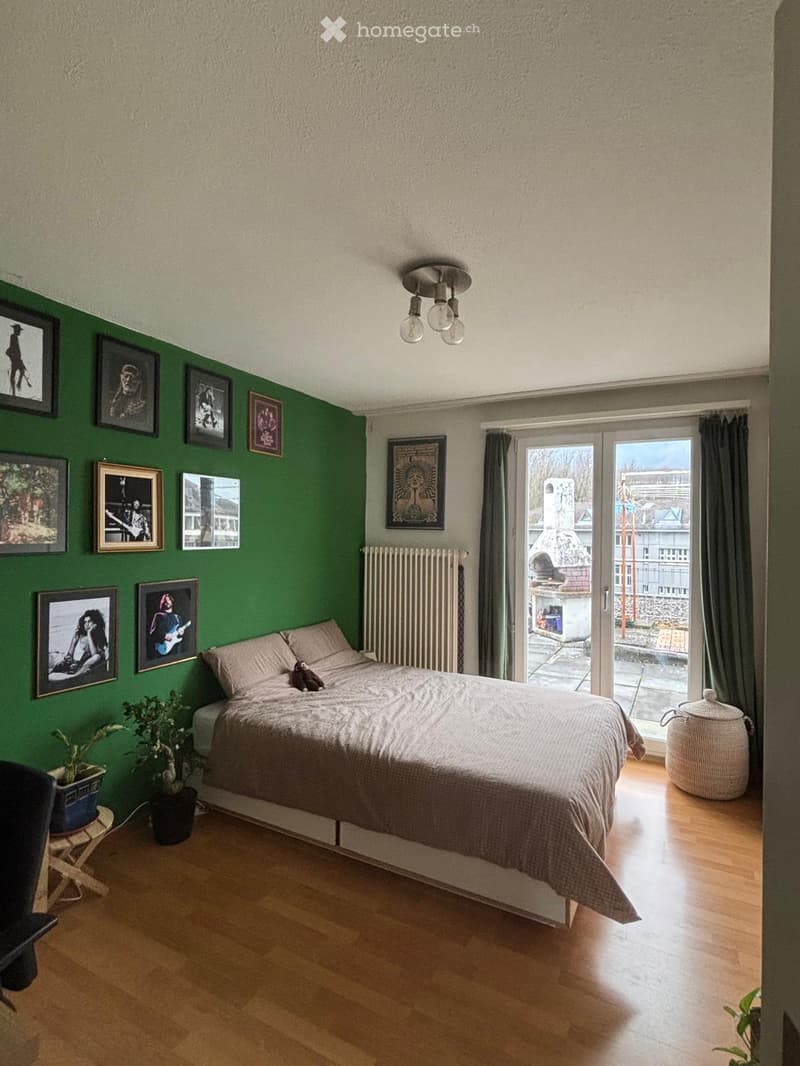 A modern bedroom with a large bed, wooden flooring, green accent wall, and several framed pictures. Features include a radiator, a small stool with a potted plant, and a large window with a view of the city.