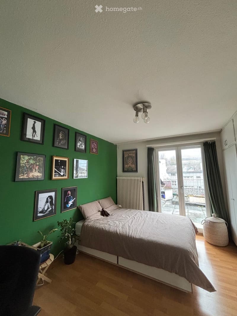 Bedroom with green accent wall, wooden floor, bed with beige bedding, framed pictures, potted plants, and a sliding glass door leading to a balcony.