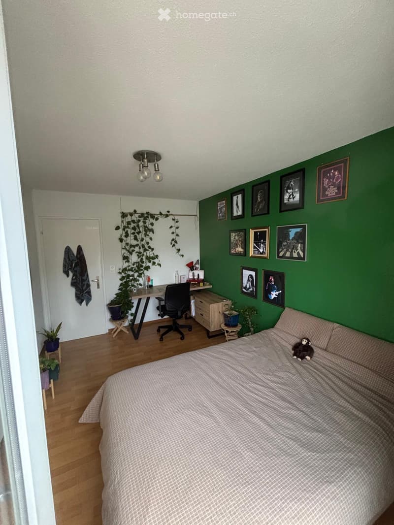 Cozy bedroom with a bed, desk, chair, and several framed pictures on the wall. Wooden floor and green accent wall.