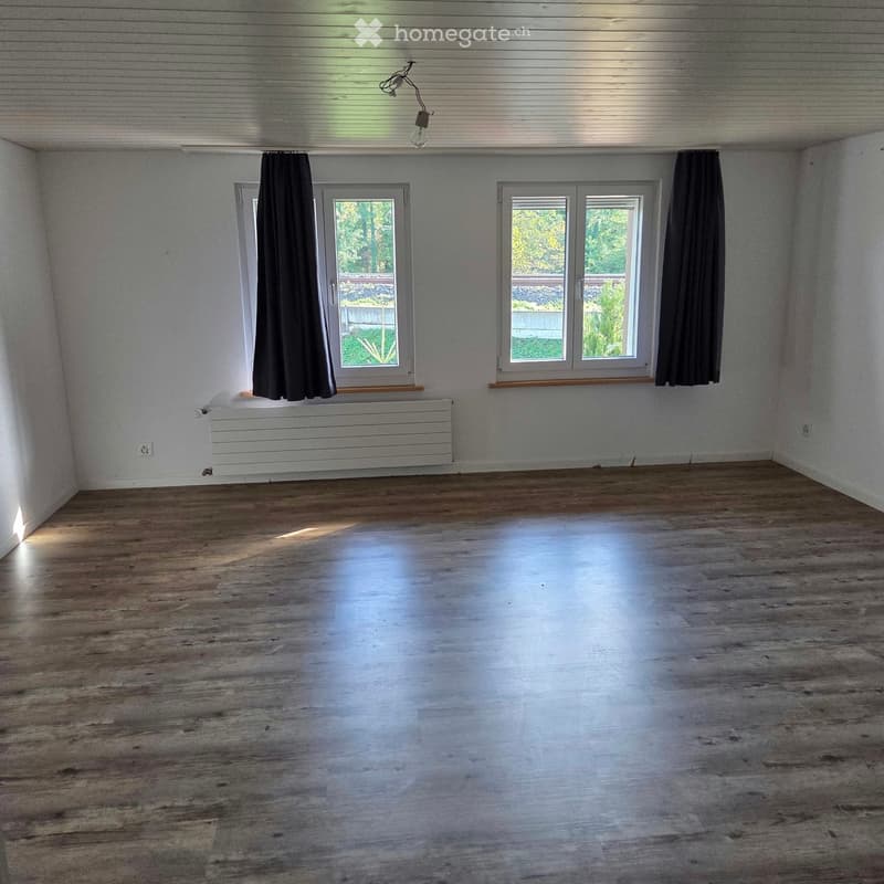 Empty room with wooden flooring, white walls, and large windows with dark curtains. A radiator is visible under one window.