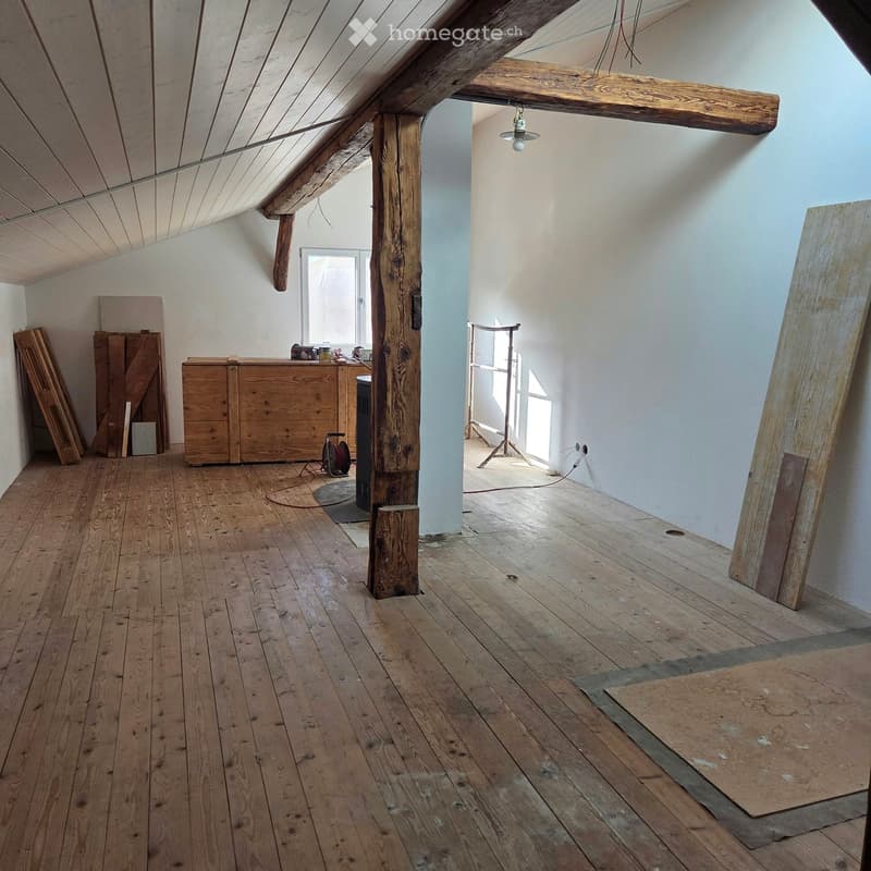 An empty attic space with wooden beams, exposed wooden floor, and a small window letting in natural light. The room features a wooden cabinet and some stacked wooden planks against the wall.