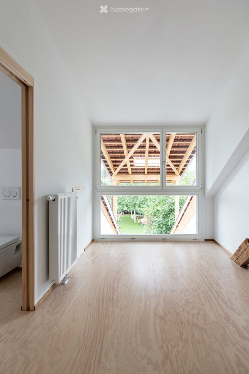 Empty room with light wooden flooring, white walls, large windows overlooking a wooden deck and garden, radiator on the left wall, and a thermostat above it.