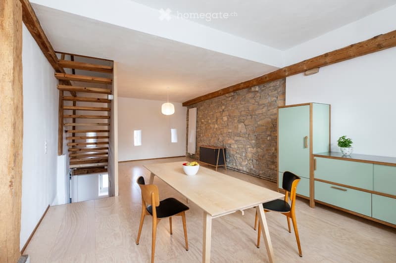 Bright, airy room with wooden flooring, exposed wooden beams, and a stone wall. Features a dining table with chairs, a light green cabinet, a small stool, and a bowl of fruit on the table.