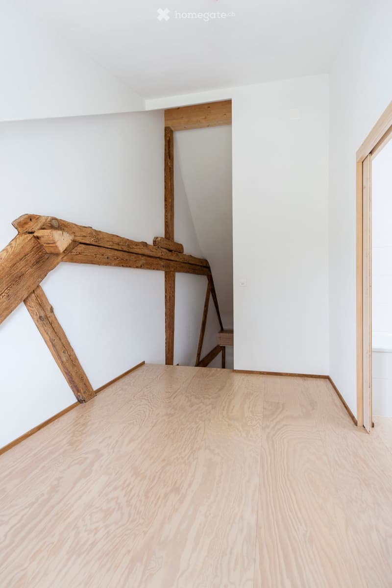 Empty room with light wooden flooring and white walls, featuring exposed wooden beams and a small staircase leading upwards.