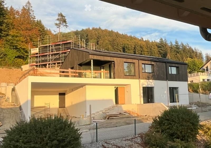 Modern two-story house under construction with a raised ground floor, garage, and large windows. The upper level features a balcony and is surrounded by trees.