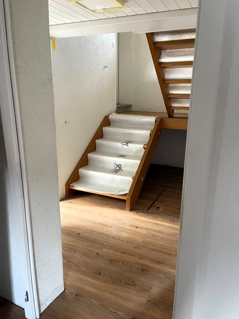Wooden staircase with white carpeted steps leading to an upper level, visible from a hallway with light wooden flooring and white walls.