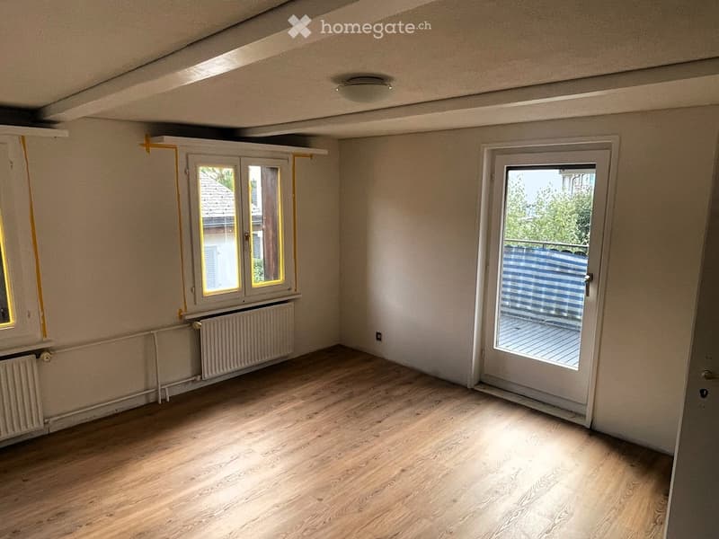 Empty room with wooden flooring, white walls, and large windows. Features a sliding glass door leading to a balcony or outdoor area.