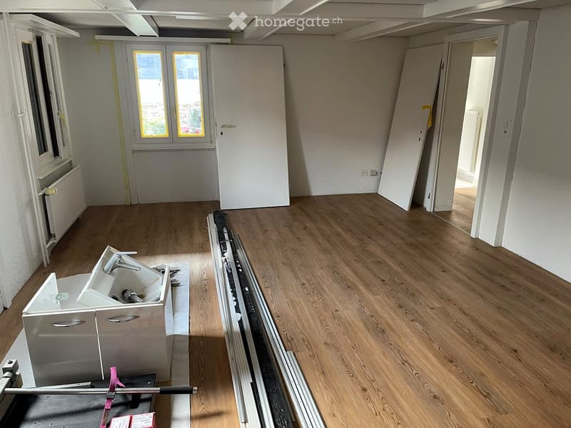 Empty room with wooden flooring, white walls, and large windows. Exposed construction materials and tools are visible, indicating ongoing renovation.
