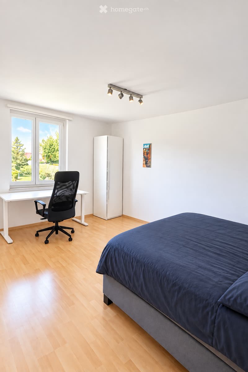 Bright bedroom with wooden flooring, large window, bed with dark blue bedding, desk with chair, and a white wardrobe.