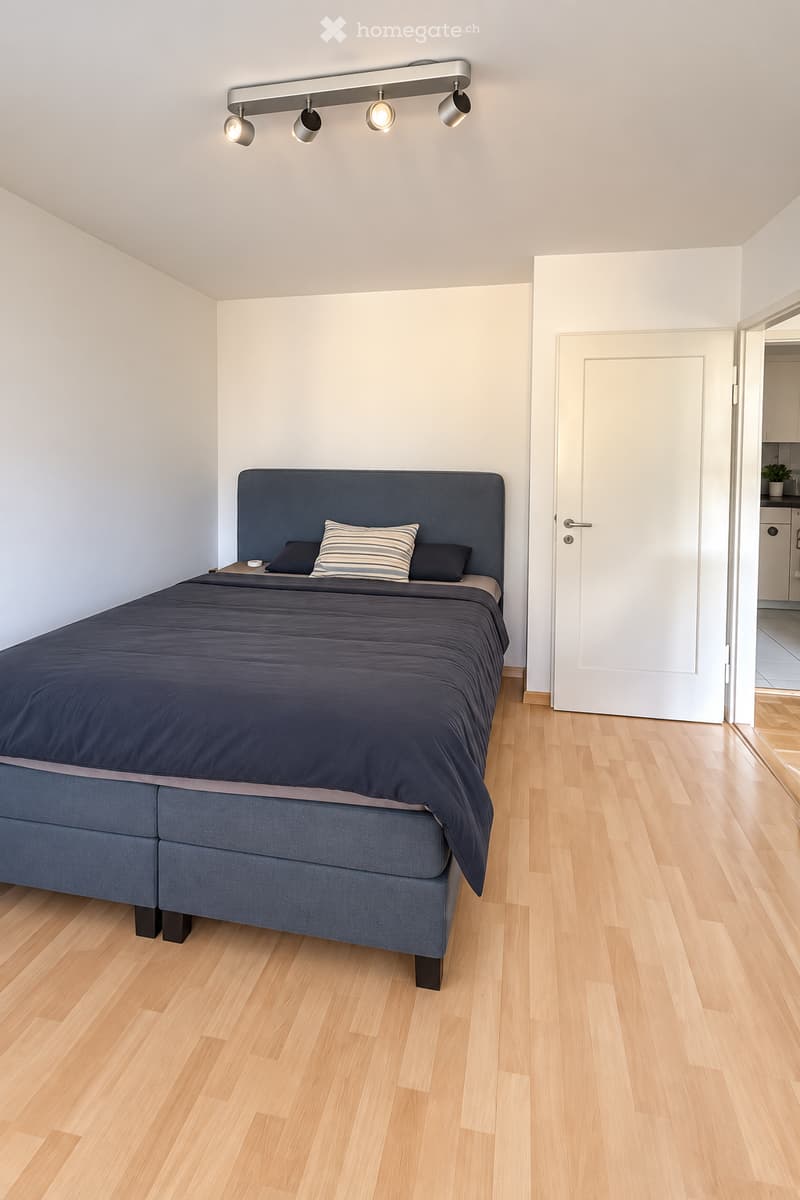 Modern bedroom with a large bed, dark blue bedding, and a nightstand. The room features light wooden flooring and white walls, with a door leading to another room visible in the background.
