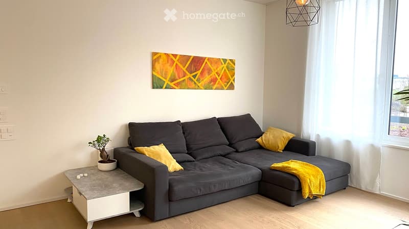 Modern living room with a grey sectional sofa, yellow throw pillows, a grey coffee table, and a small potted plant. Large windows with white curtains and a colorful abstract painting on the wall.