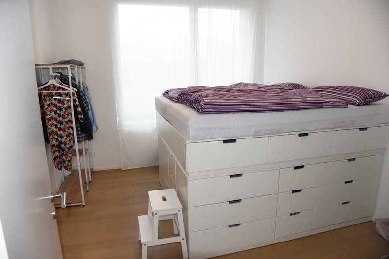 Bright bedroom with a lofted bed on a white drawer unit, a small white stool, a clothing rack with hanging clothes, and natural light from large windows.