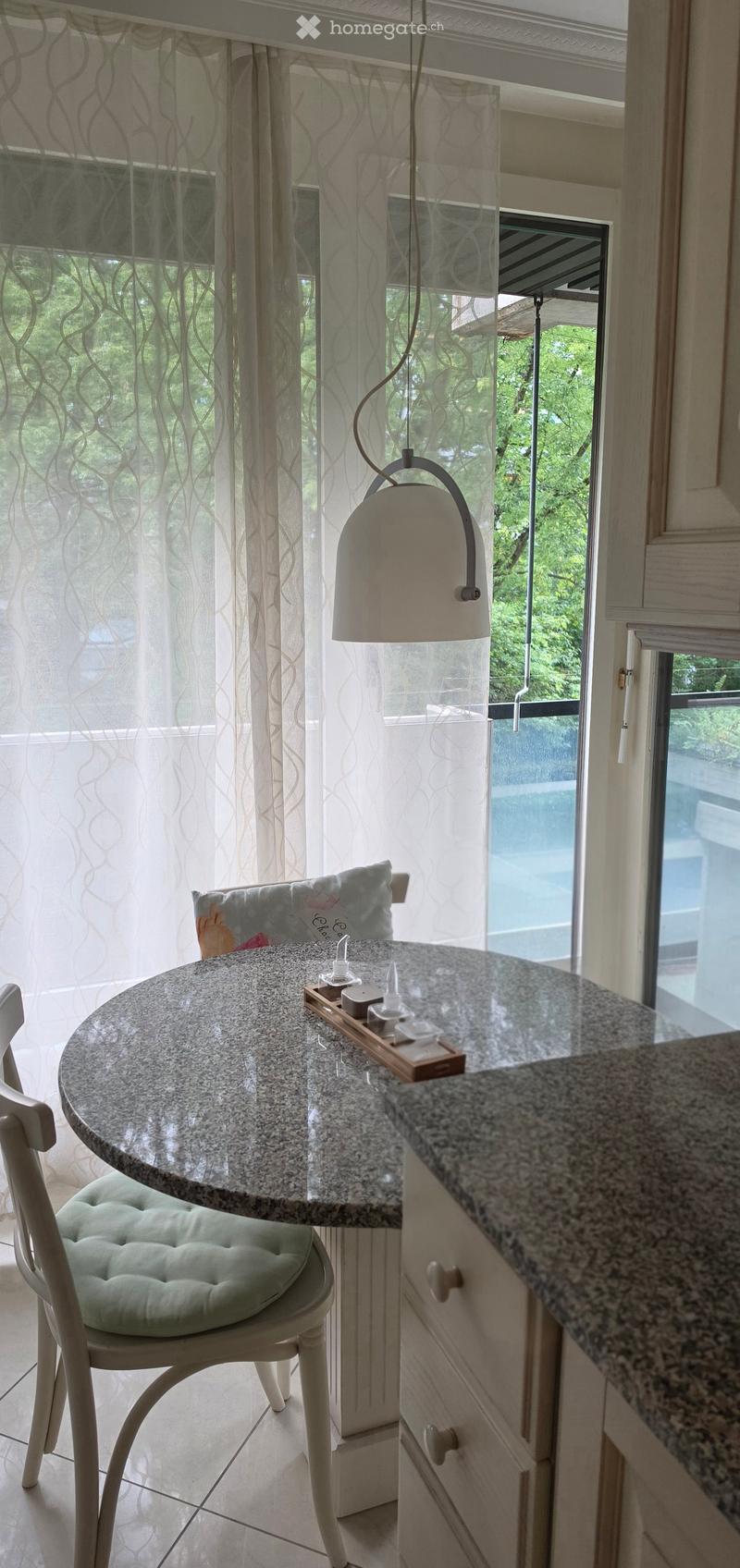 A small, well-lit room with a round table, a single chair, a cabinet, and a hanging lamp. The room has large windows with sheer curtains, offering a view of greenery outside.
