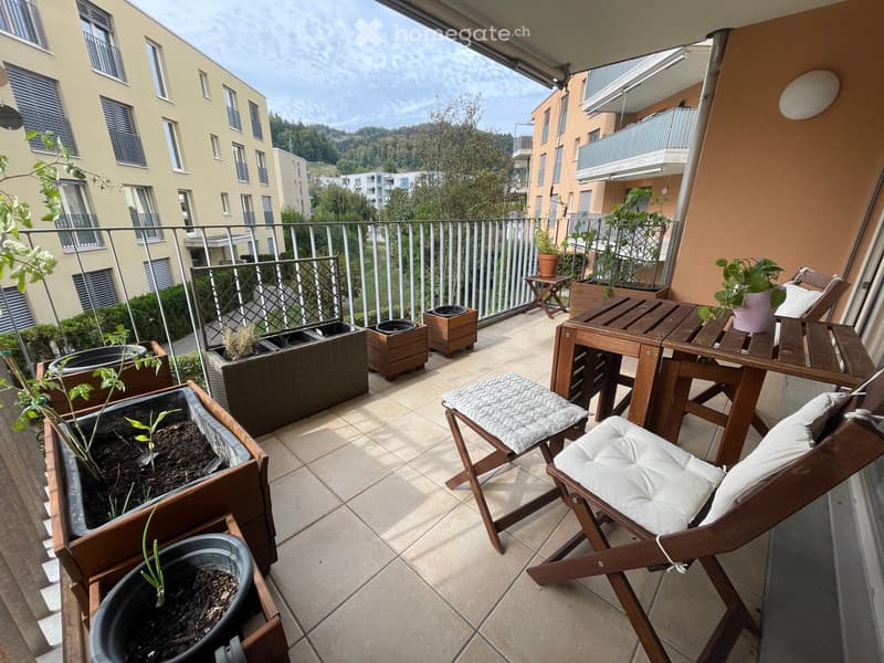 Balcony with wooden table, chairs, potted plants, and a view of the surrounding buildings and greenery.