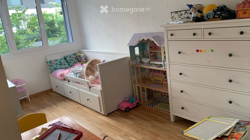 A cozy children's bedroom with a built-in sofa bed, a dollhouse, a toy car, a small chair, a table with books and a picture frame, and a large drawer unit.