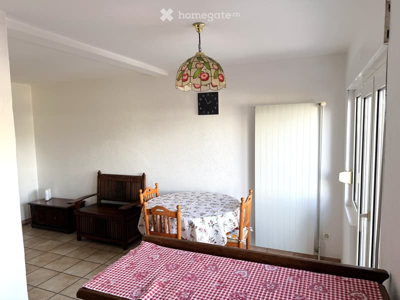 A furnished room with a wooden bench, a table with a floral tablecloth, wooden chairs, a clock, a radiator, and a decorative lamp.