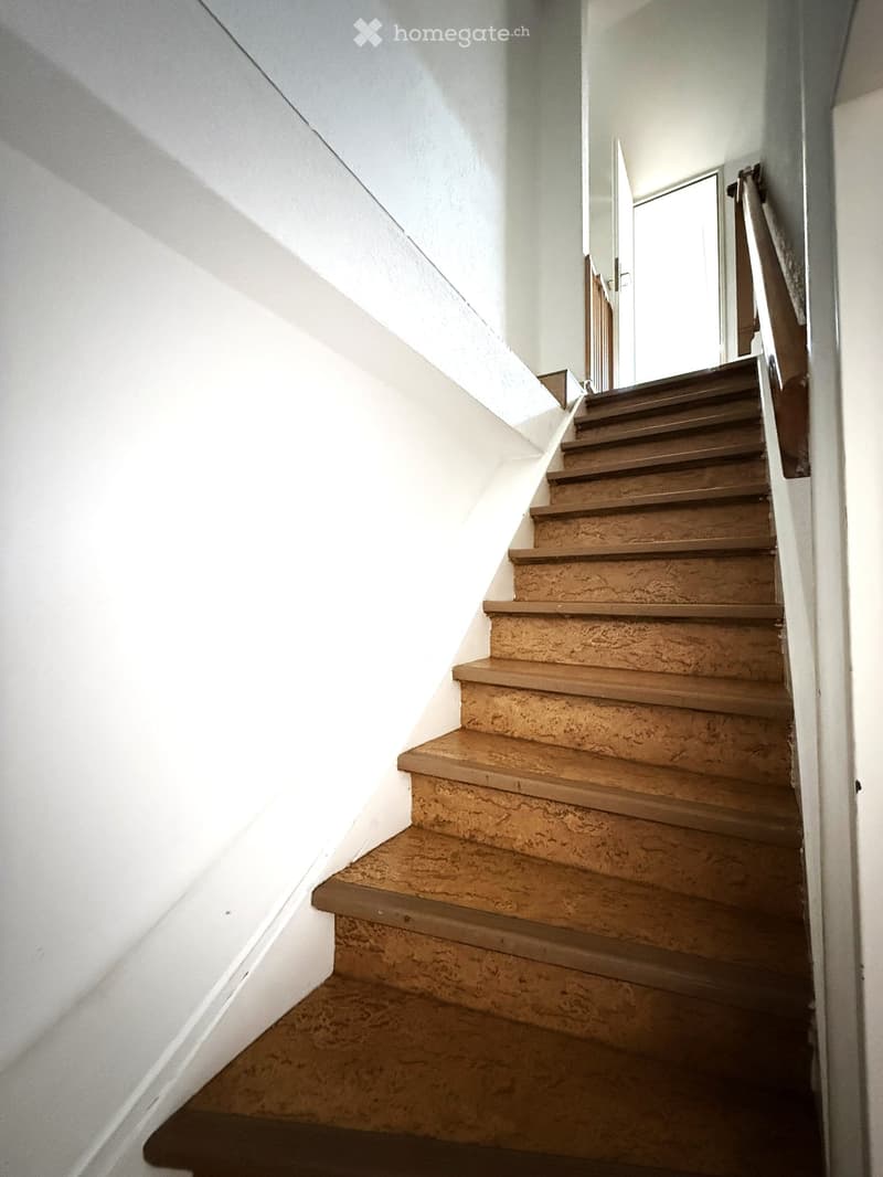 A narrow staircase with wooden steps and a metal handrail, leading upwards in a well-lit interior space with white walls.