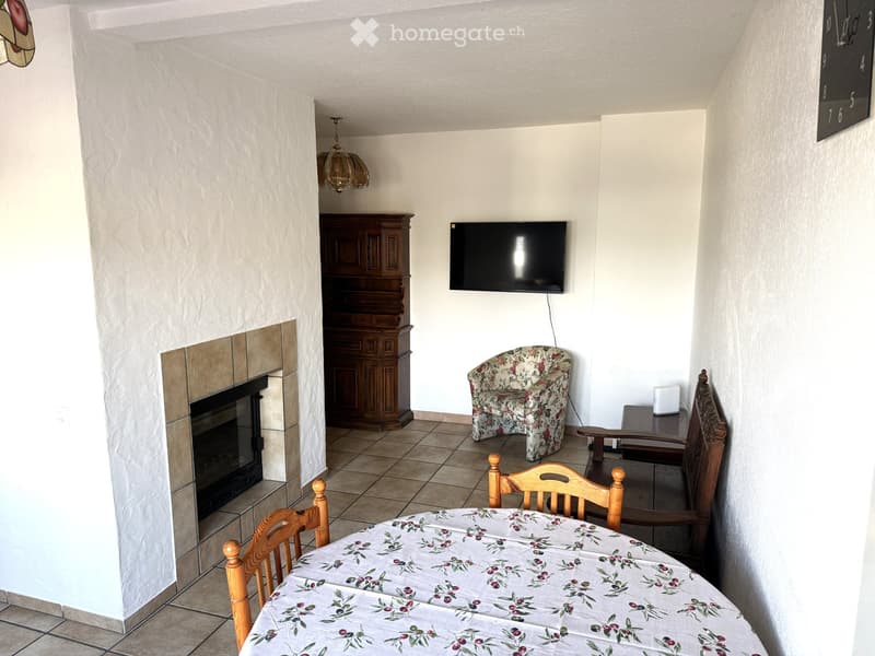 Living room with a fireplace, television, wooden dining table, chairs, floral-patterned sofa, and a wooden cabinet.