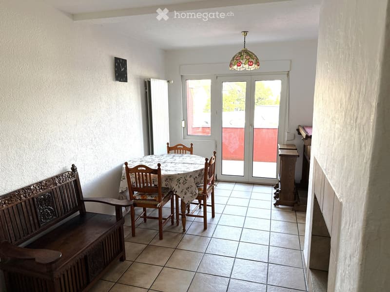 Dining area with a round table, four chairs, a wooden bench, a decorative clock, and a stained-glass lamp hanging from the ceiling. The room has tiled flooring and large glass doors leading to a balcony.