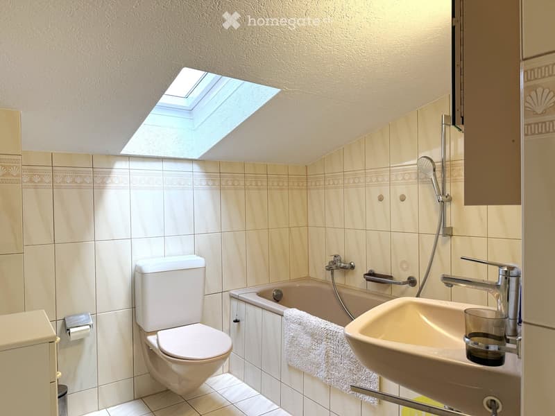 Bathroom with a toilet, bathtub, sink, and a towel on the edge of the tub. The room features beige tiles and a skylight window above the bathtub.