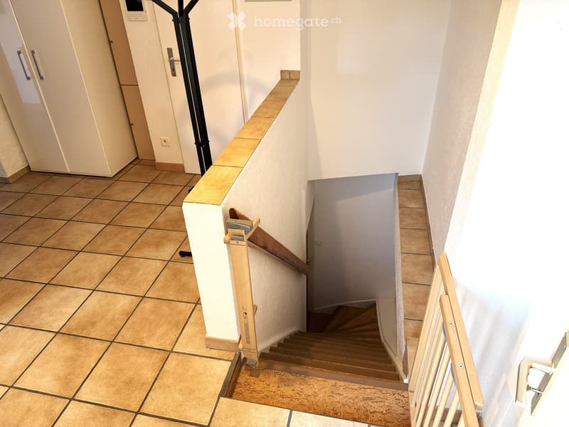 A modern hallway with tiled flooring, a wooden staircase with a handrail, and a small wooden ramp for accessibility.