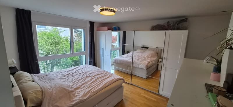 Bright bedroom with wooden flooring, large window with view, double bed with beige bedding, mirrored wardrobe, nightstand, and lamp.