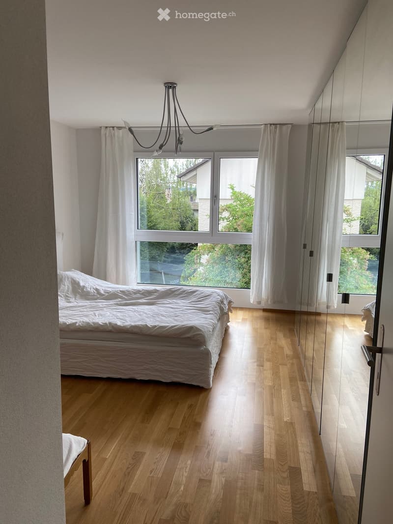 Bright bedroom with wooden flooring, large window with white curtains, and a neatly made bed.