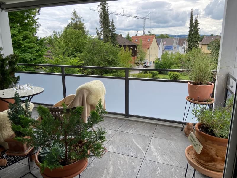 Balcony with tiled floor, metal railing, potted plants, small table, and a blanket draped over a chair.