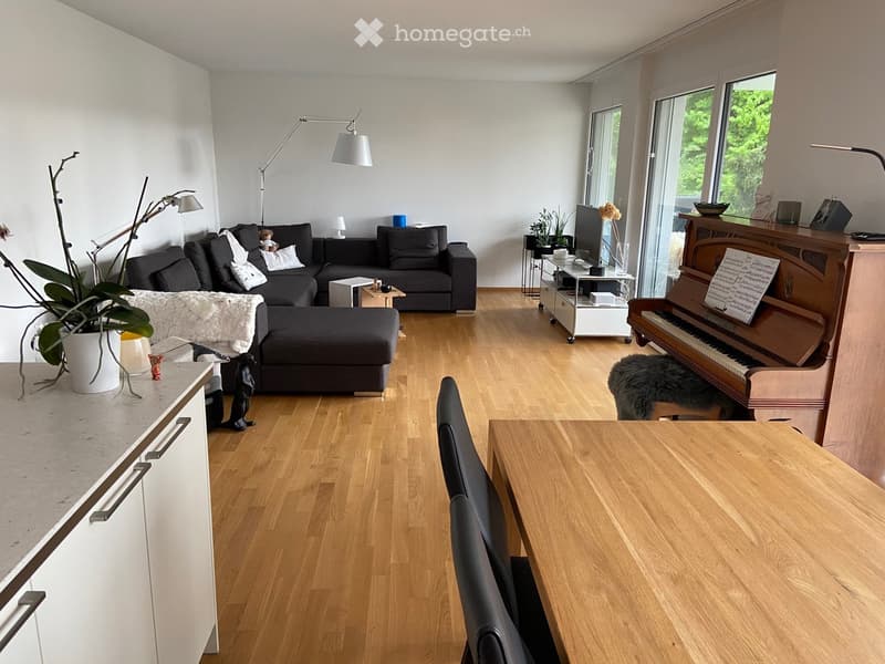 Modern living room with wooden flooring, large windows, a black sectional sofa, a wooden piano, a dining table with chairs, and decorative plants.