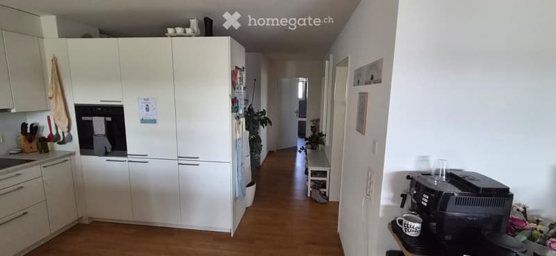 A well-lit hallway with wooden flooring, leading to a kitchen area with white cabinets, a sink, and various kitchen utensils. There is a coffee machine on a small table with a mug and a vase of flowers nearby.