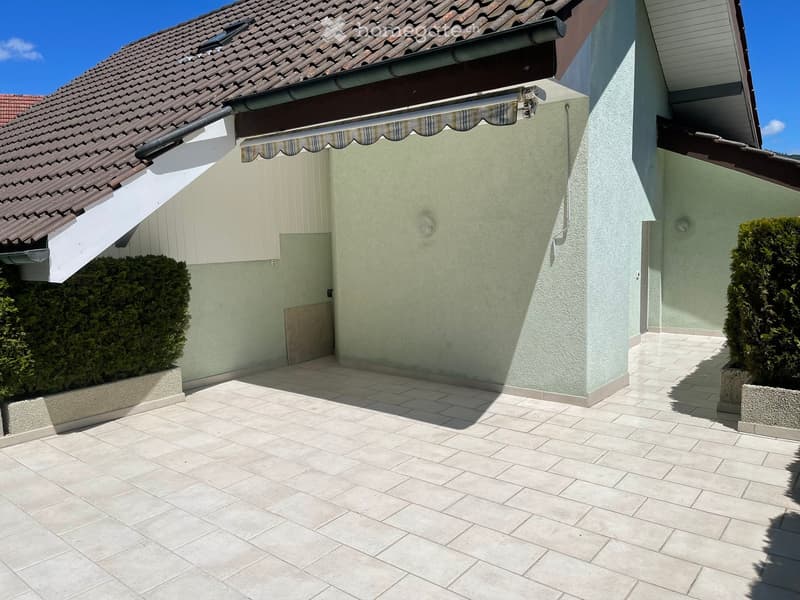 A tiled patio area adjacent to a house with light green walls, a tiled roof, and a retractable awning. Two neatly trimmed bushes flank the entrance.