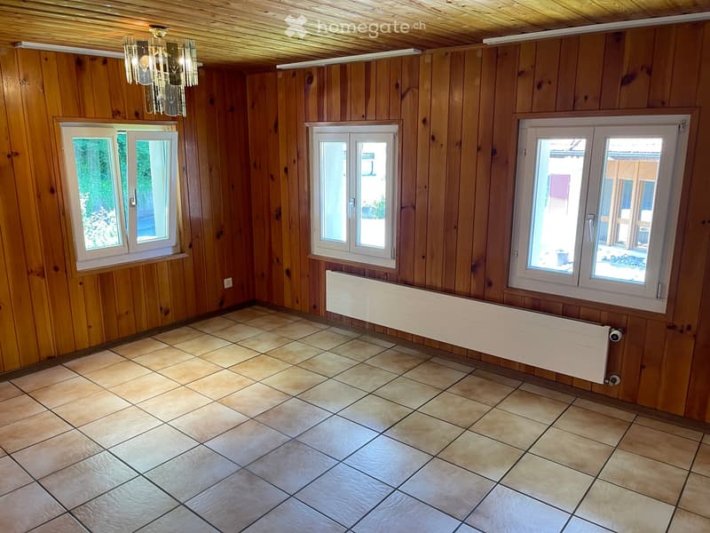 Empty room with wooden walls, tiled floor, three windows, and a radiator. No furniture or equipment present.