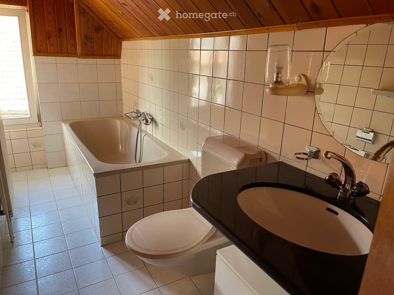 Bathroom with a bathtub, toilet, sink, and mirror. The walls and floor are tiled, and there is a window on the left side.