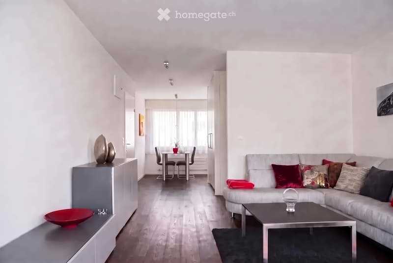 Modern living and dining area with white walls, wooden flooring, a grey sofa with colorful cushions, a coffee table, a side table with decorative items, and a dining table with chairs.