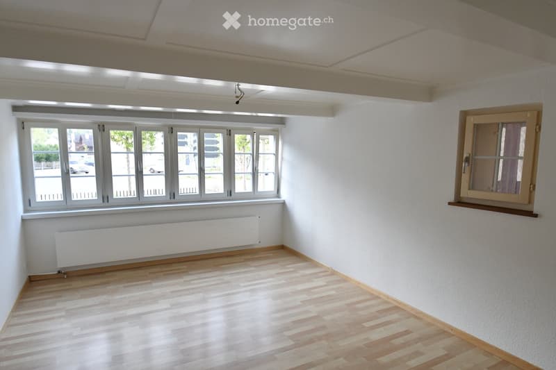 Empty room with wooden flooring, white walls, large windows, radiator, and a small wooden shelf on the right side.