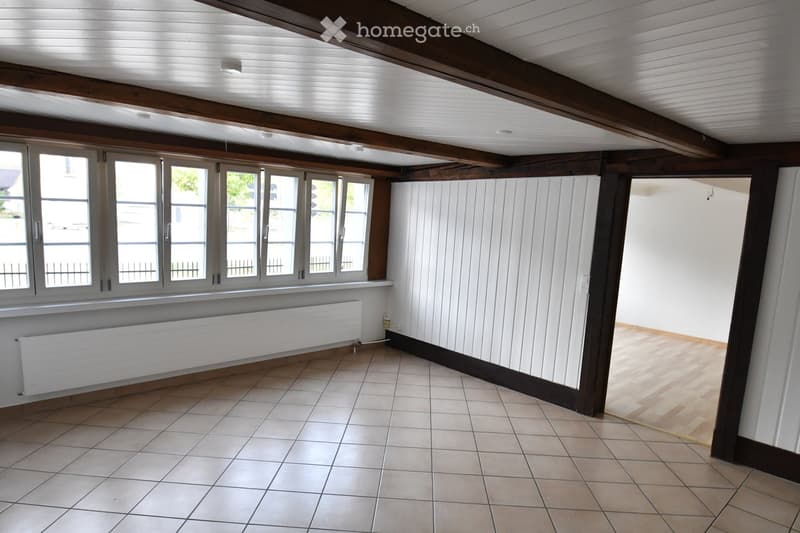 Empty room with exposed wooden beams, tiled floor, large windows, radiator, and white paneled walls.