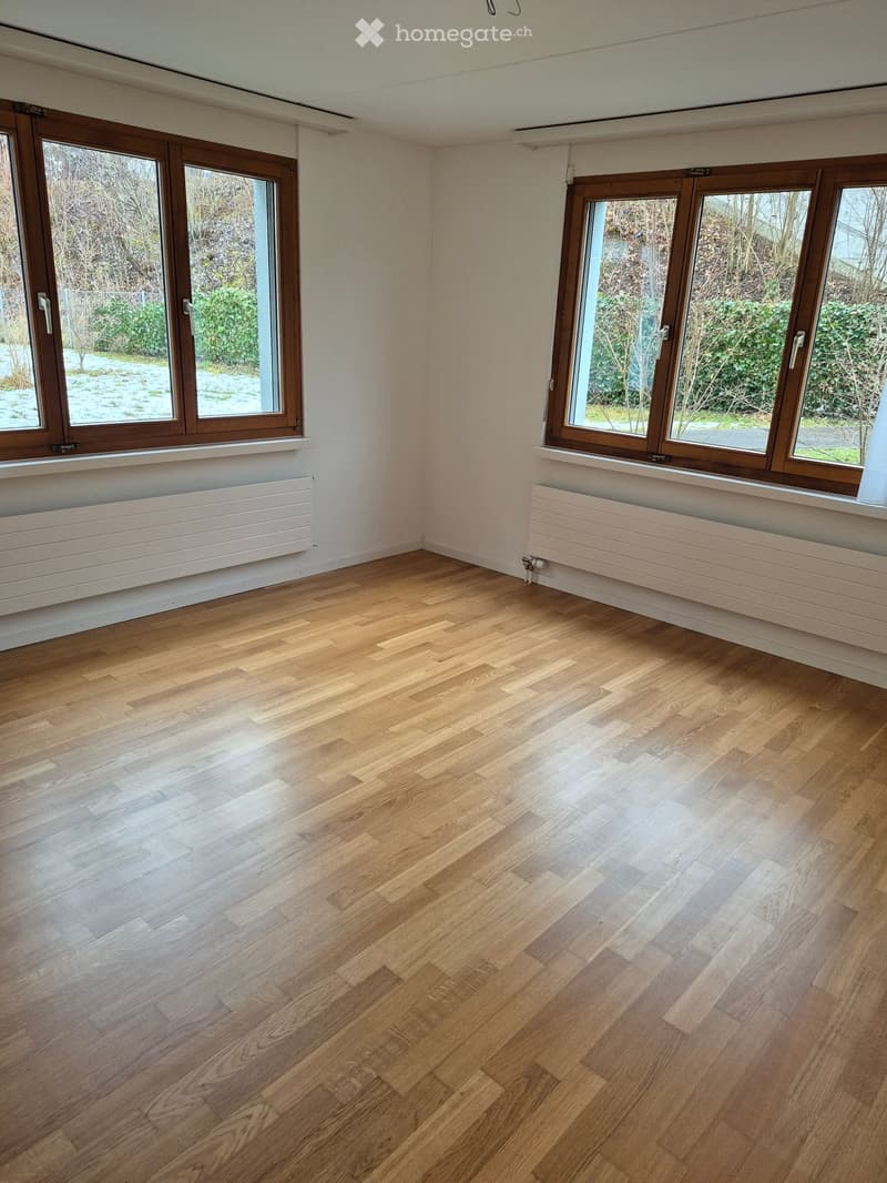 Empty room with wooden flooring, white walls, large windows with wooden frames, and radiators beneath the windows.