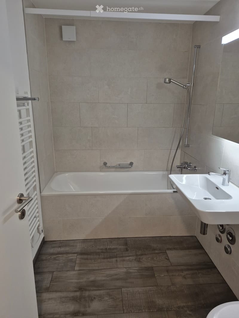 Modern bathroom with a bathtub, sink, and a handheld showerhead. Light-colored tiles and wooden flooring.