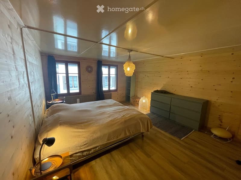 Cozy bedroom with wooden walls and floor, large windows, a bed with white bedding, a nightstand with a lamp, a drawer unit, and a hanging pendant light.