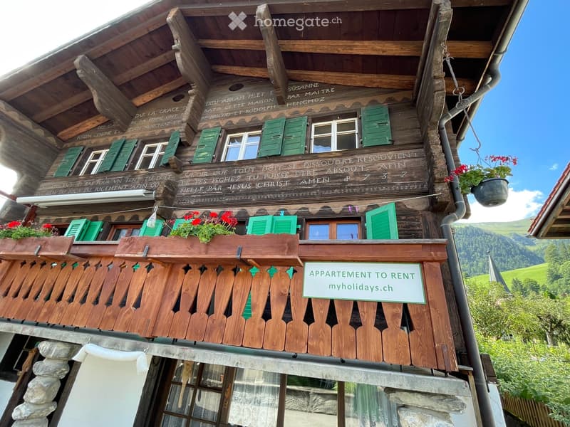 Traditional wooden chalet with green shutters, balconies, and a sign advertising rental apartments. The building features intricate wooden carvings and a stone base.